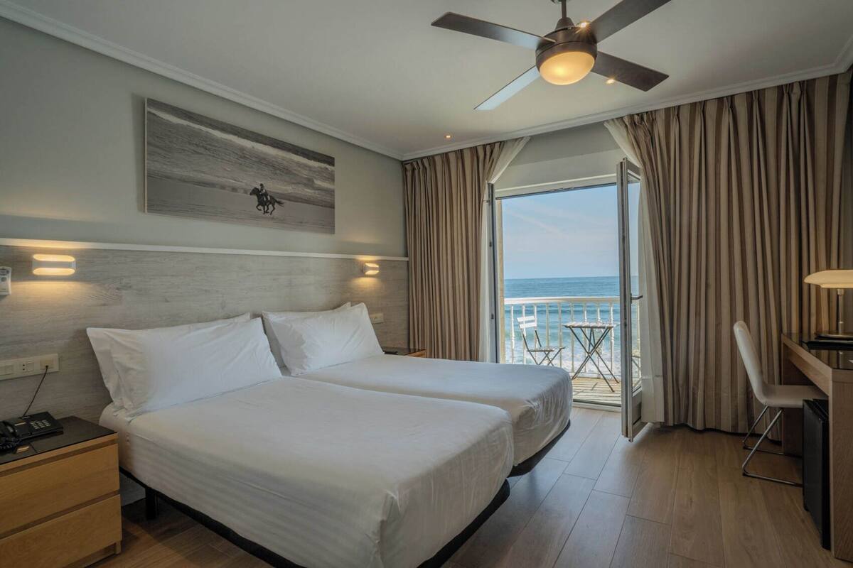 Habitación de hotel con dos camas, ventilador de techo, escritorio con silla, balcón con vista al mar y un cuadro en la pared.