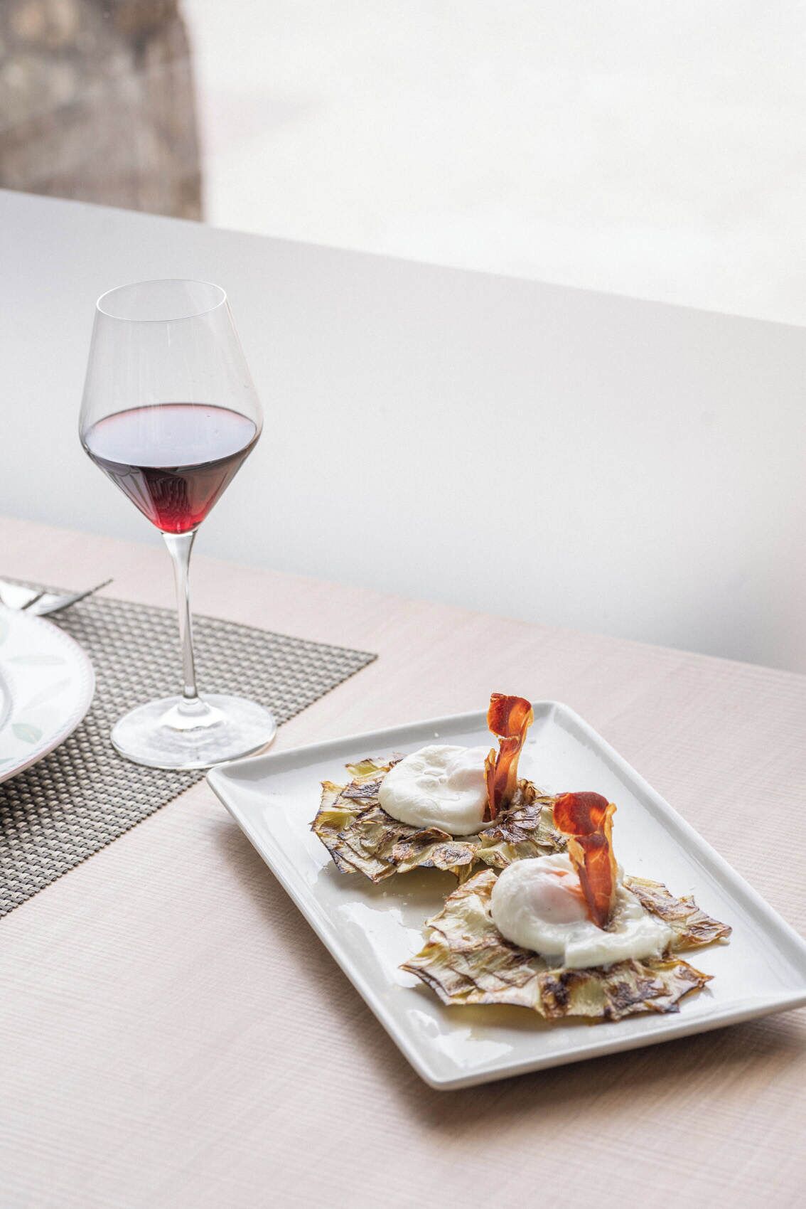 A dining table with a white plate of food, a glass of red wine, and a woven placemat.