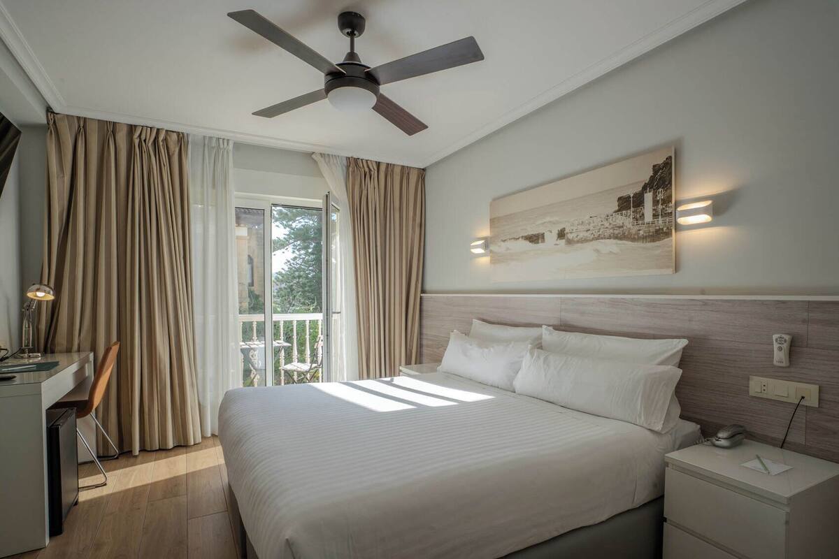 Ein Hotelzimmer mit Bett, Schreibtisch, Stuhl, Deckenventilator, einem Balkon mit Vorhängen und einer wandmontierten Leuchte.