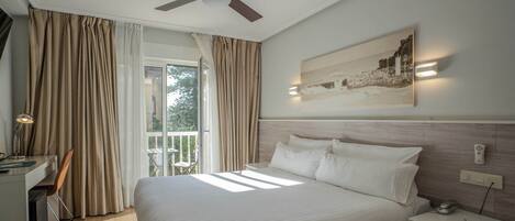 A hotel room with a bed, a desk, a chair, a ceiling fan, a balcony with curtains, and a wall-mounted light fixture.