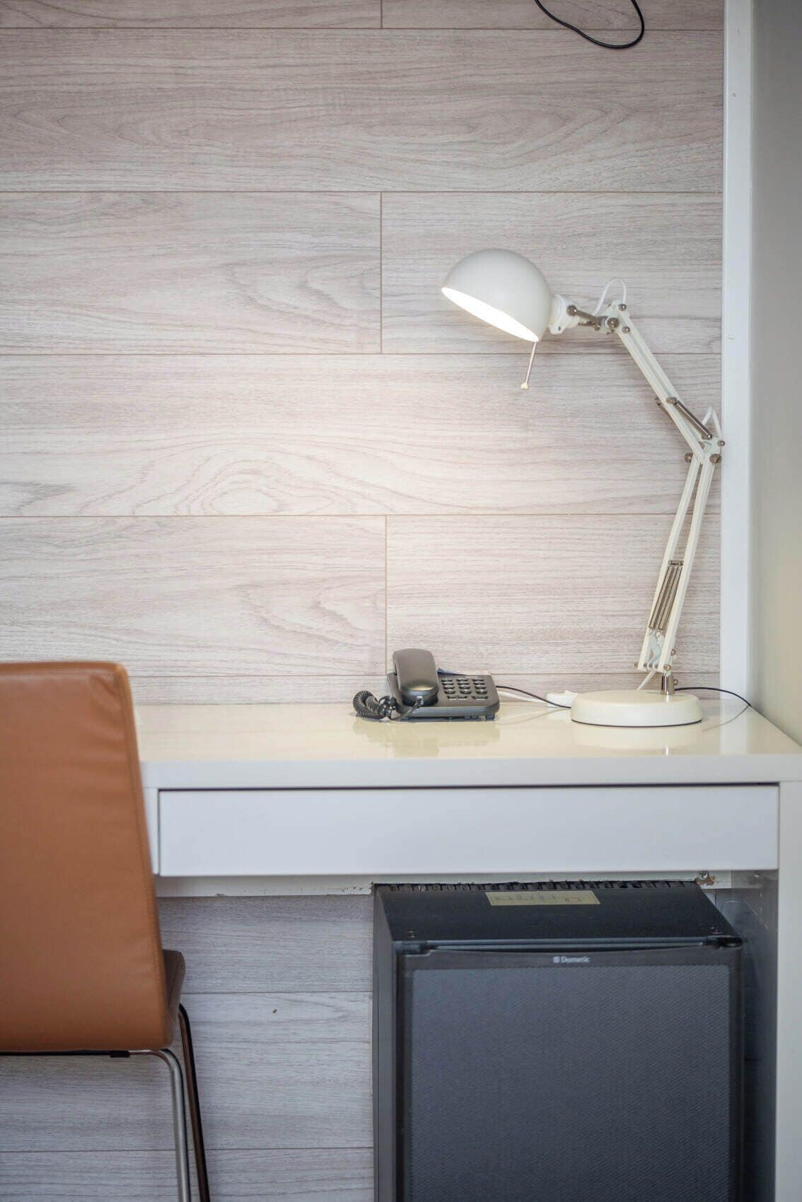 Escritorio con lámpara, teléfono y un pequeño dispositivo negro, posiblemente una impresora, frente a una pared de madera.