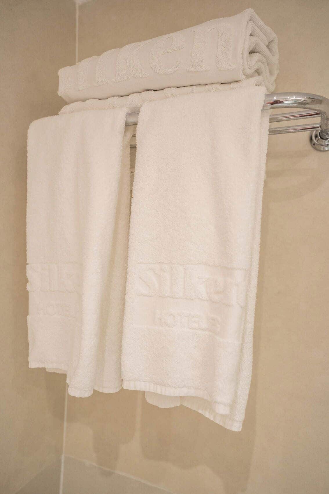A set of white towels with embossed text, hanging on a metal towel rack in a bathroom with beige tiles.