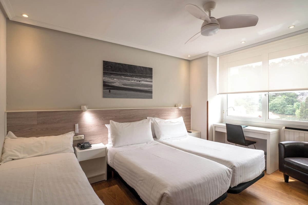Ein Hotelzimmer mit zwei Betten, einem Schreibtisch, einem Stuhl, einem Deckenventilator und einem großen Fenster.