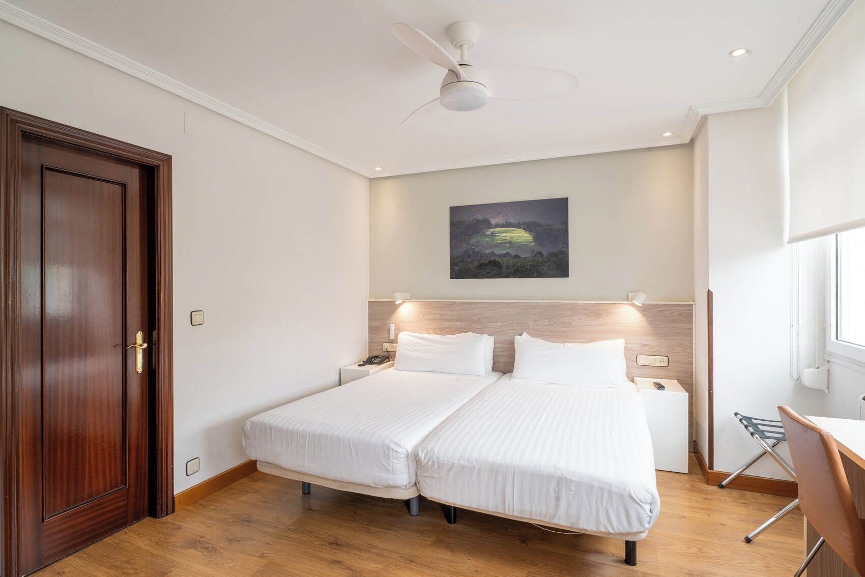 A hotel room with two beds, a wooden door, a ceiling fan, a painting on the wall, and a window.