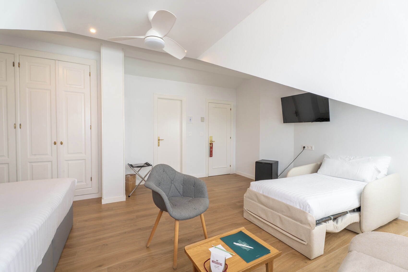 A bedroom with a bed, a chair, a table, a TV, and a ceiling fan.