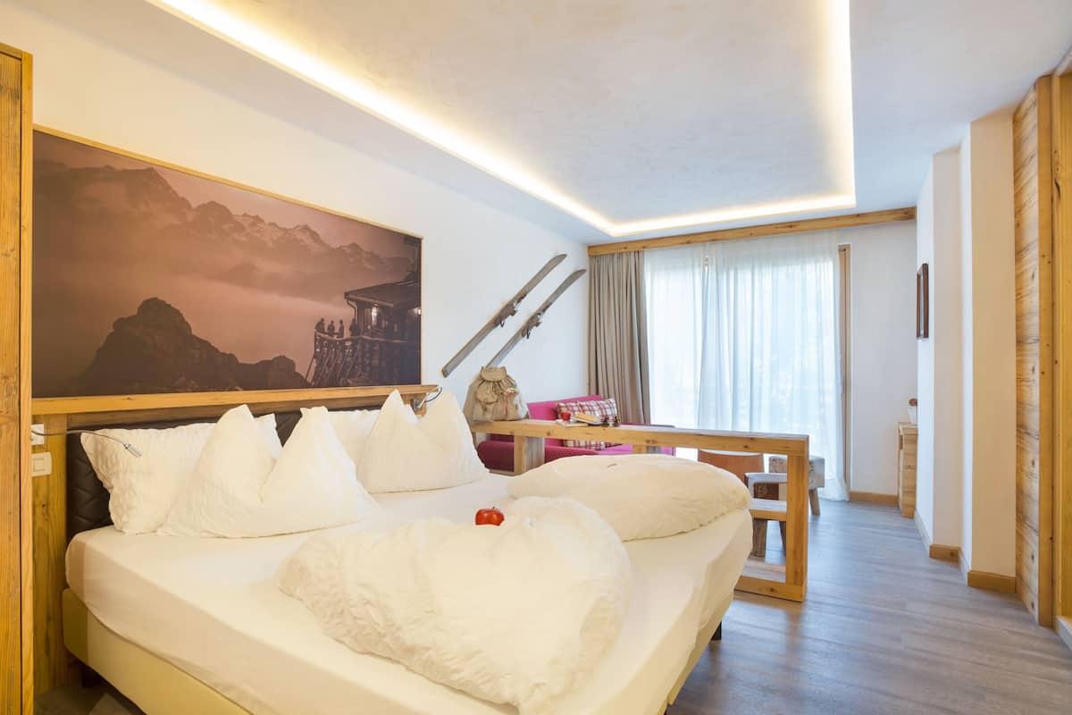 A hotel room with a large bed, a wooden headboard, a painting of mountains, and a window with curtains.