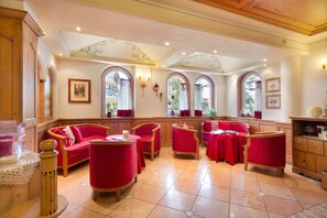 Un comedor con sillas tapizadas en rojo y mesas, armarios de madera y ventanas arqueadas con cortinas.