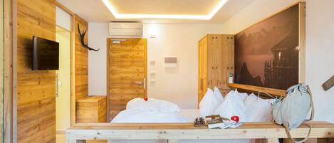 A hotel room with a wooden bed, a mounted TV, a painting on the wall, and a wooden desk with a bag and books.