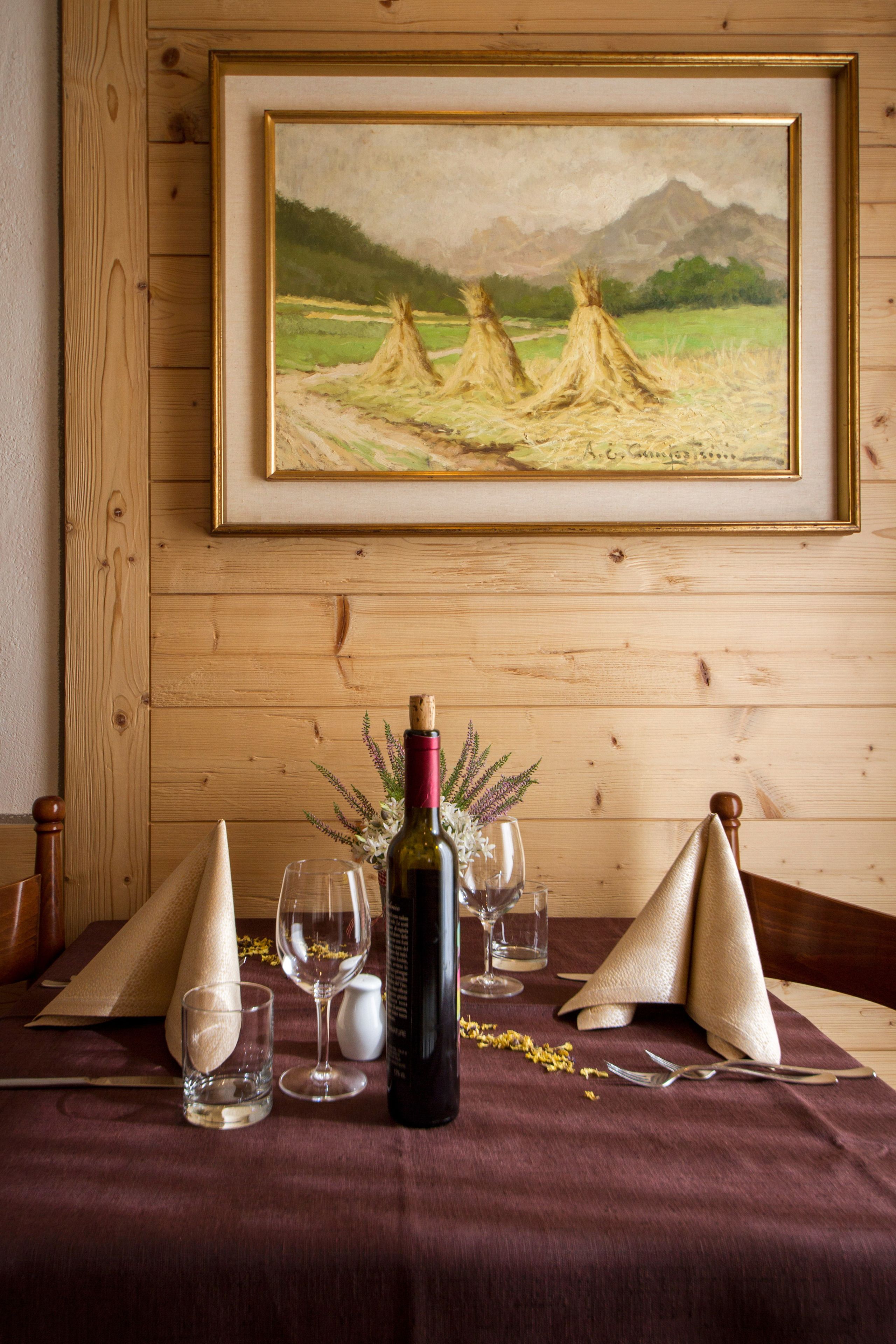 A dining table with a wine bottle, glasses, and a painting of a landscape with mountains.