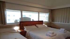 A hotel room with two beds, a wooden headboard, a bedside table with a lamp, and a view of buildings through large windows.