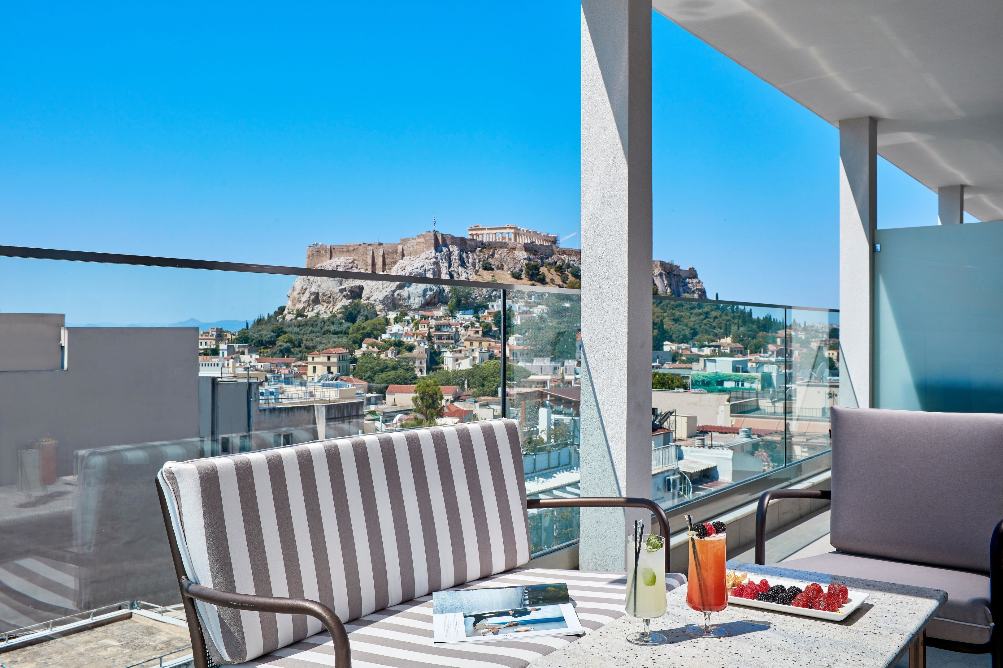 Ein Balkon mit Blick auf eine historische Burg auf einem Hügel, ein Tisch mit Getränken und Früchten sowie gestreifte Stühle.