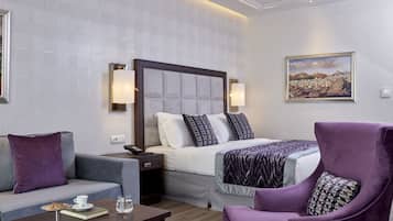 Quarto de hotel com cama, sofá, poltrona, mesa de cabeceira e um quadro na parede.
