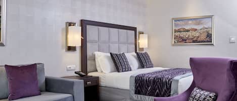 A hotel room with a bed, a sofa, a chair, a nightstand, and a painting on the wall.