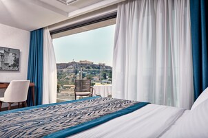 A hotel room with a large bed, a desk, and a chair. There is a window with a view of a city and a hill.
