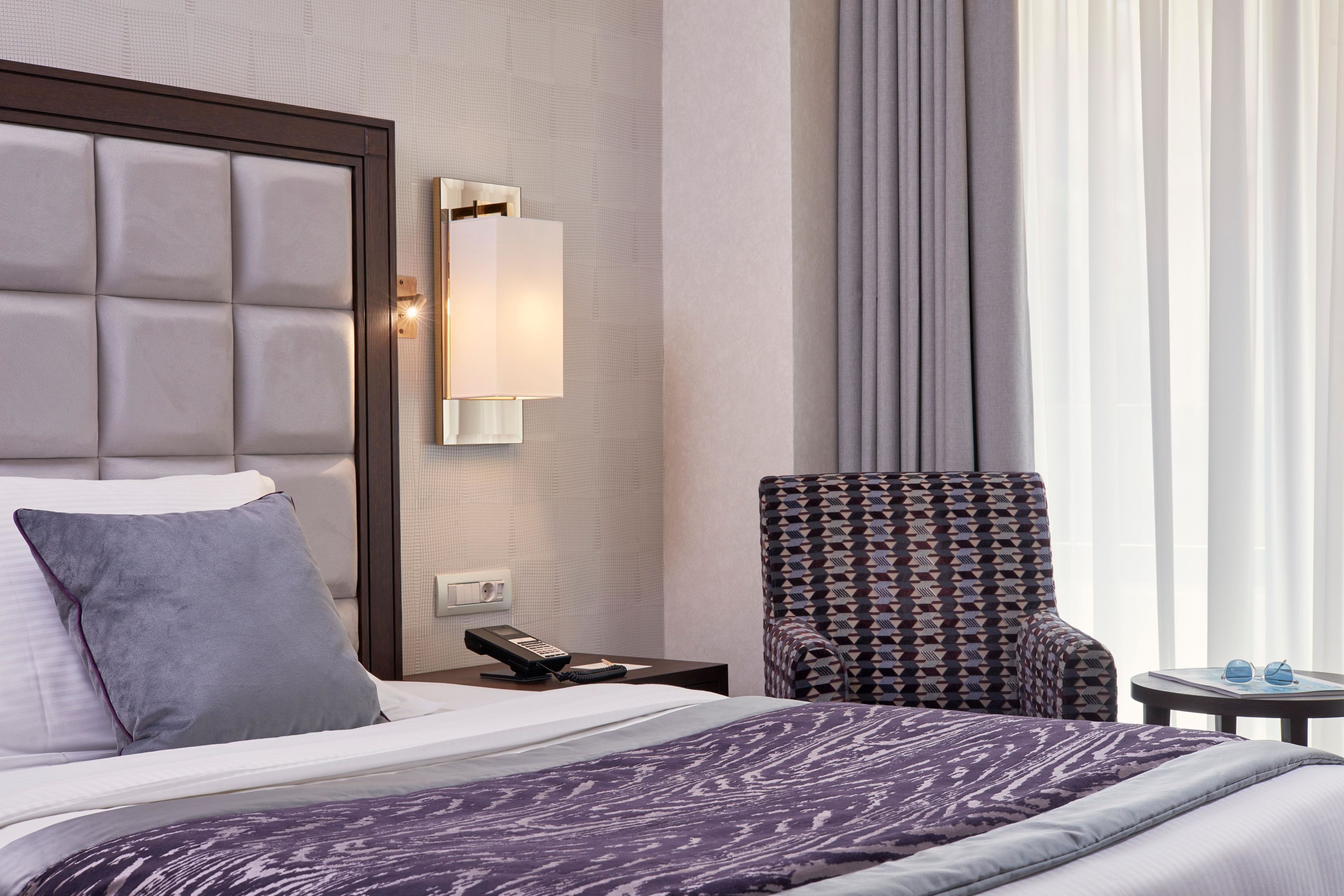 A hotel room with a bed, a nightstand with a phone, a chair, and a window with curtains.