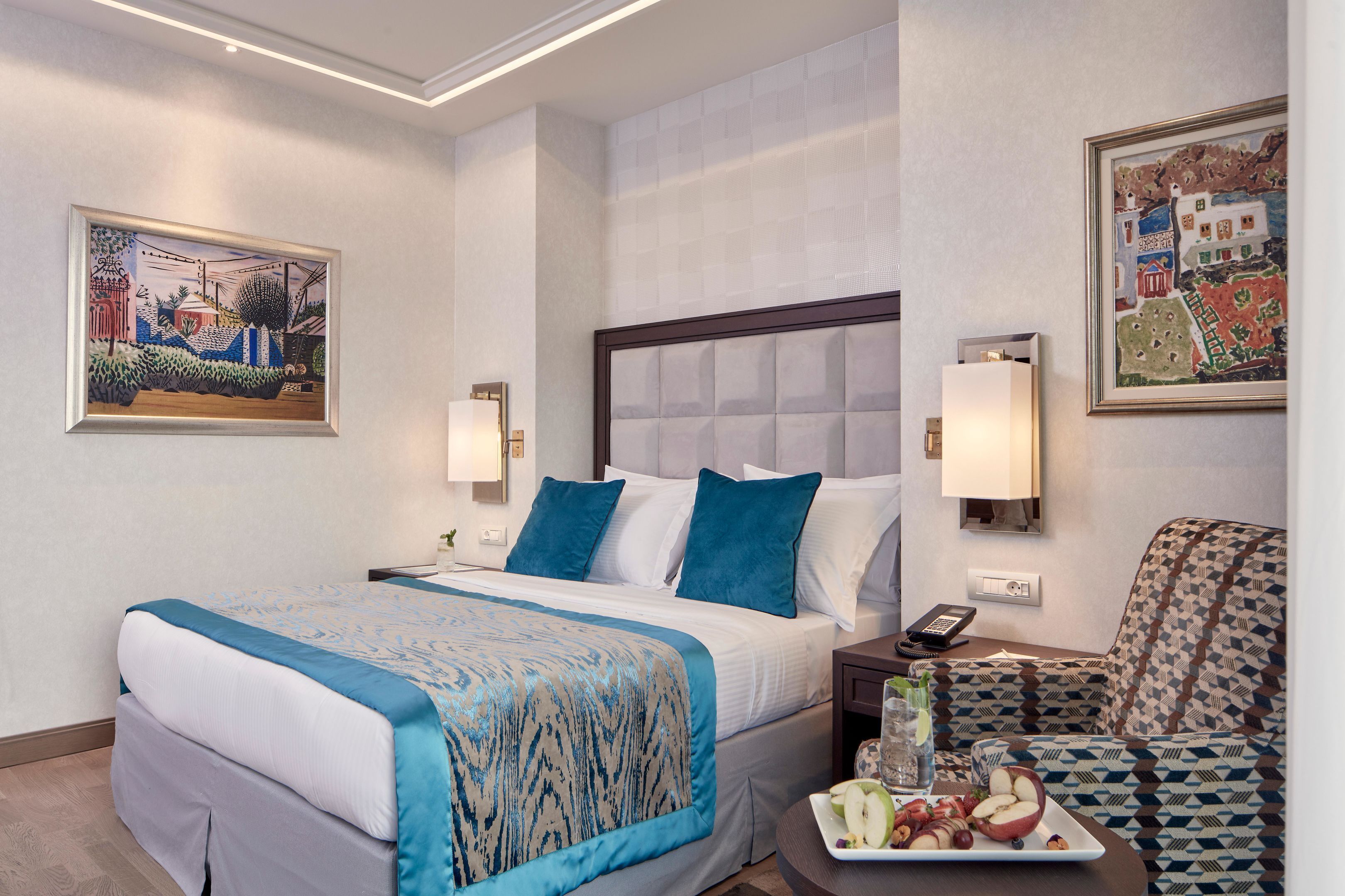 A hotel room with a bed, bedside table, lamp, artwork, and a seating area with a tray of fruit.