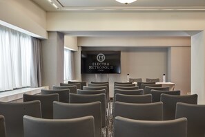A conference room with rows of chairs, a large screen displaying "ELECTRA METROPOLIS ATHENS," and a window with curtains.