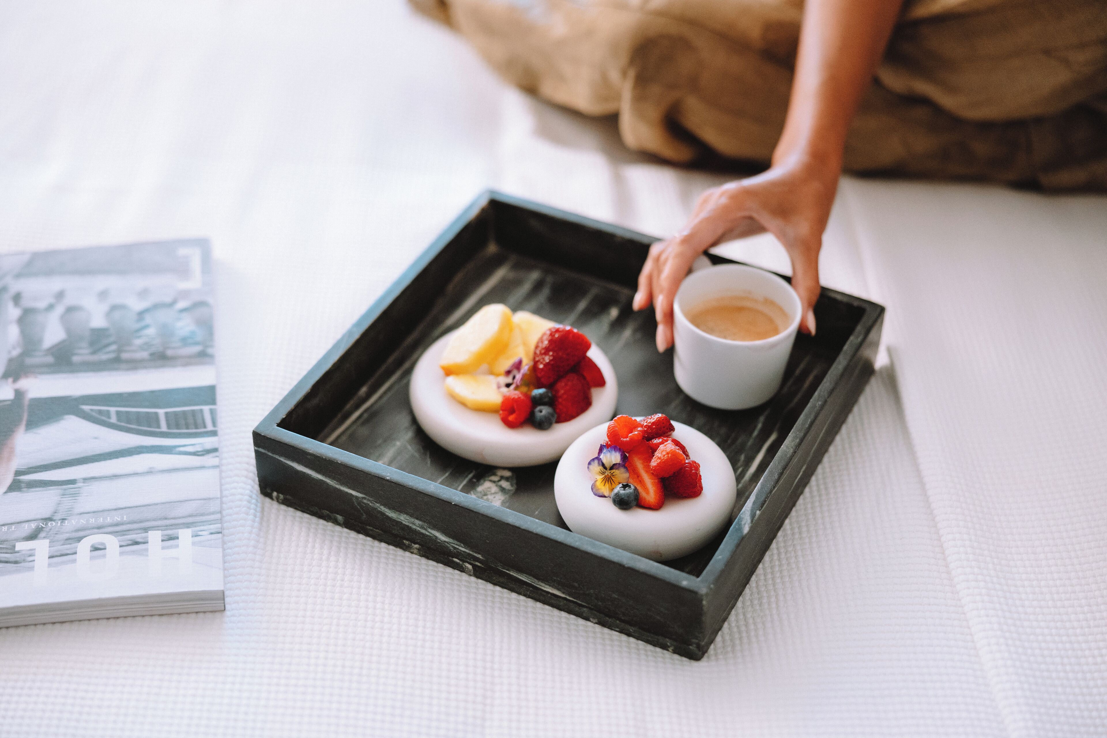 A tray with two desserts topped with fruit, a cup of coffee, and a magazine on a bed.