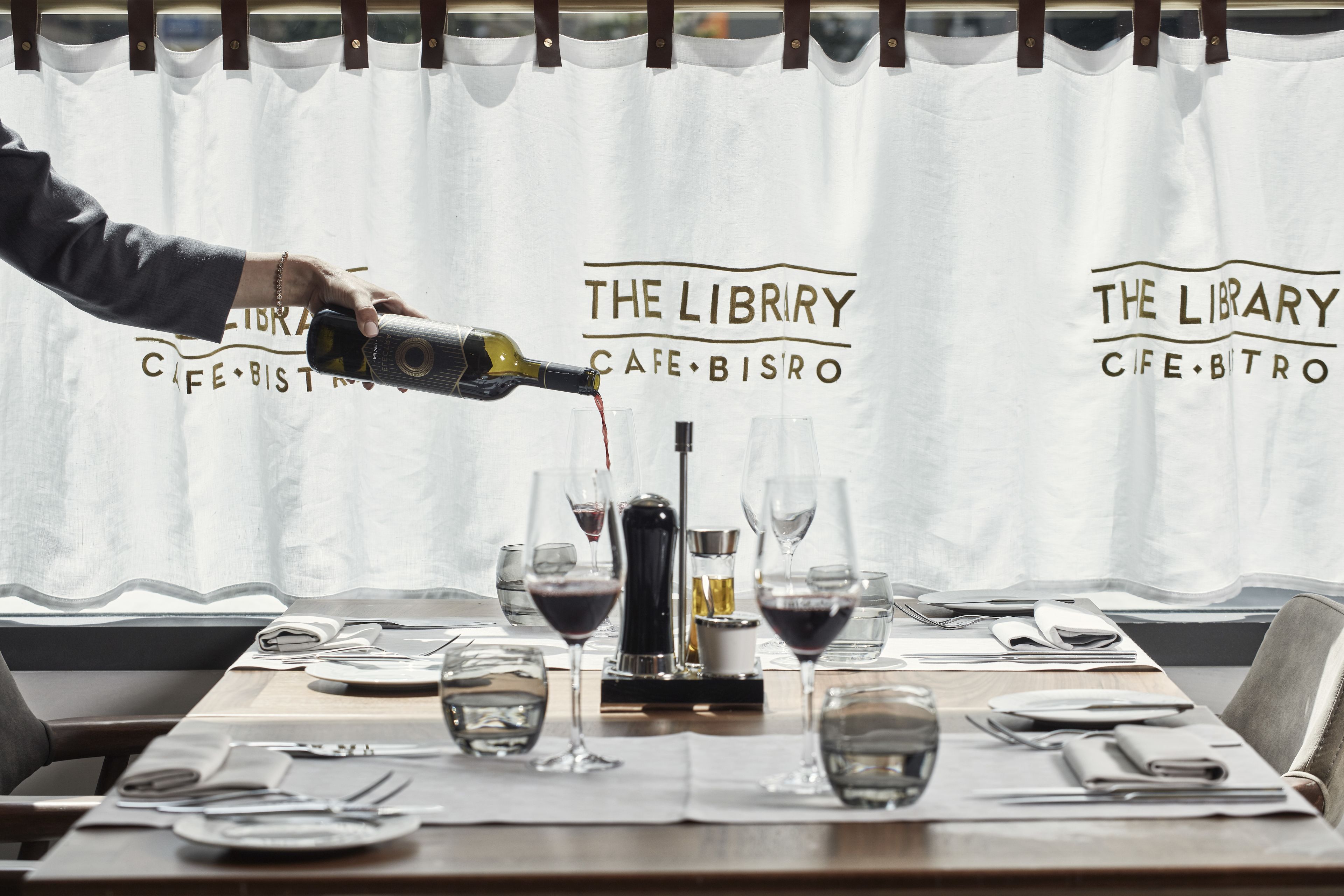 Una mesa dispuesta para comer, copas de vino, una botella de vino siendo servida y un atril para el menú.