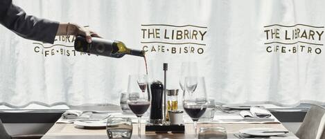 Una mesa dispuesta para comer, copas de vino, una botella de vino siendo servida y un atril para el menú.