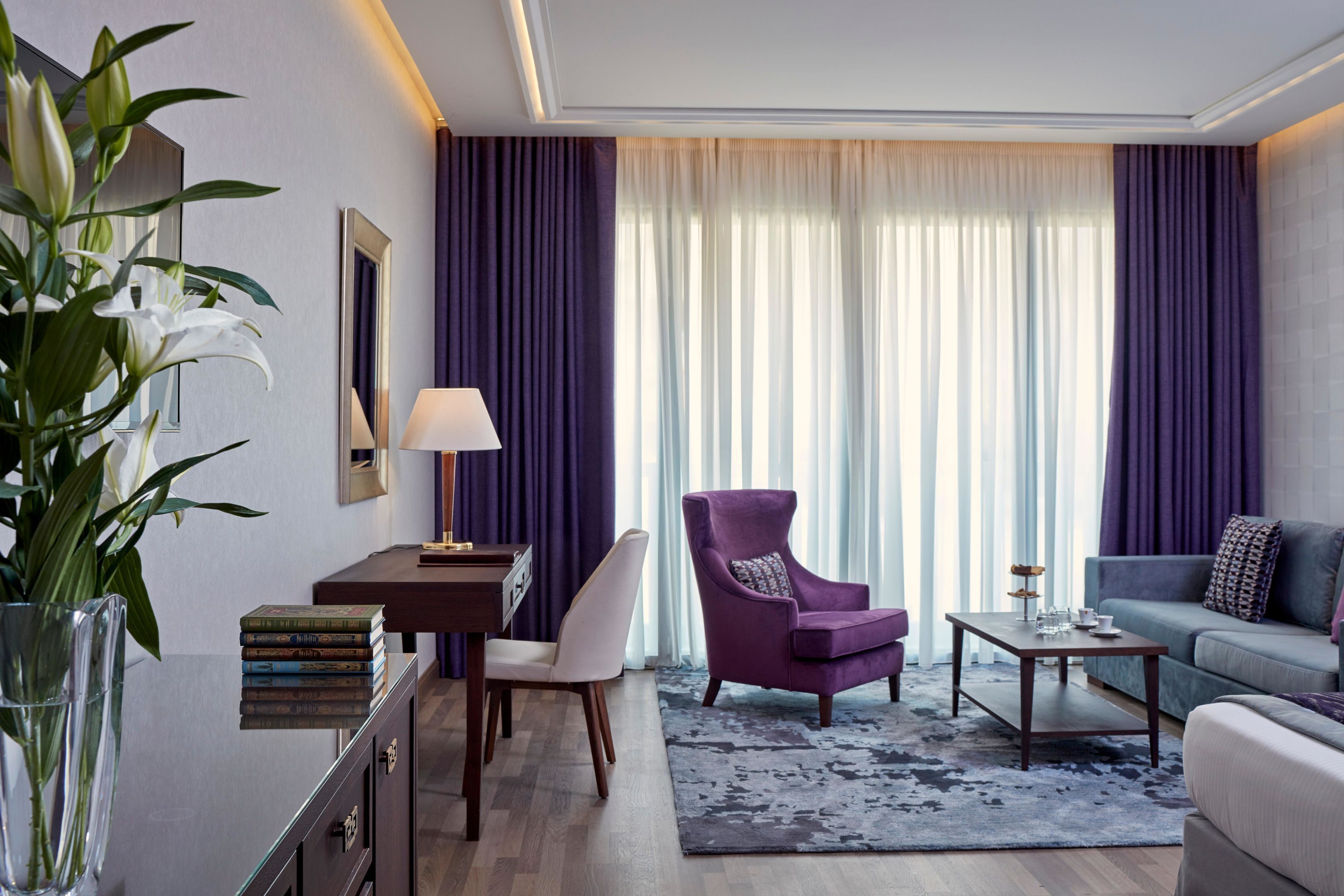 A modern hotel room with a desk, purple armchair, sofa, and a large window with curtains.