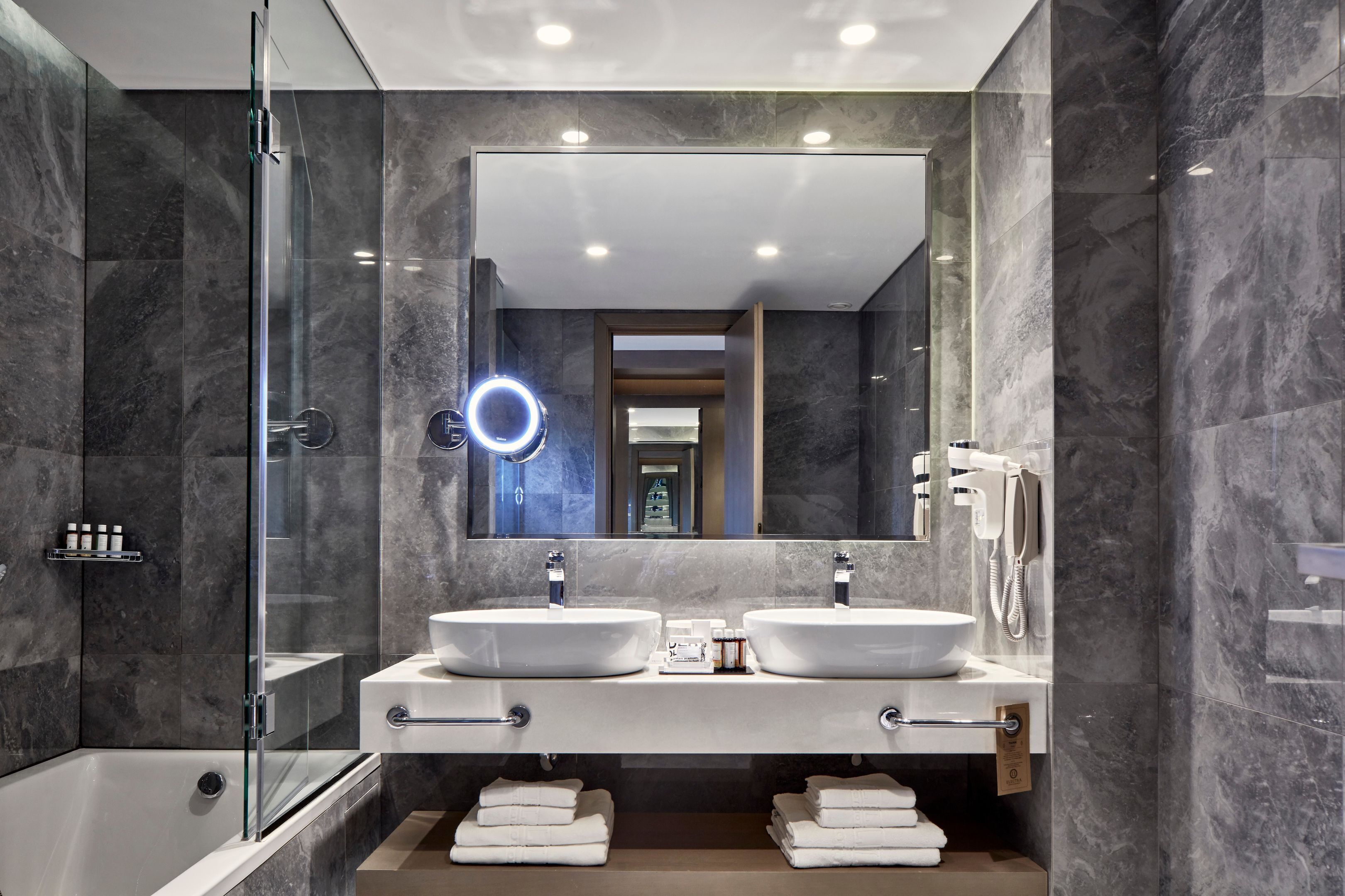 Un baño moderno con dos lavamanos, cabina de ducha con mampara de vidrio y un espejo grande con luz de aro.