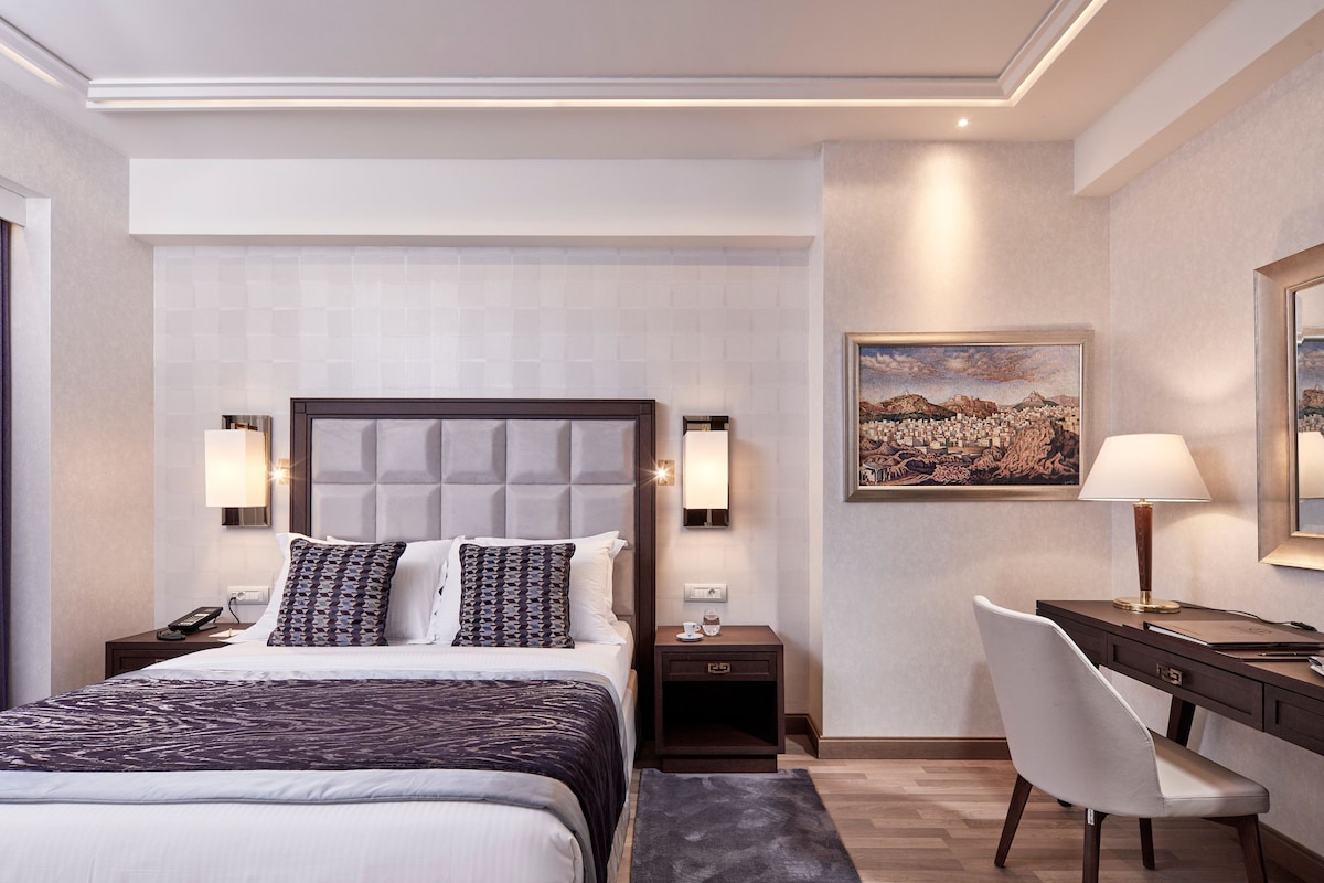A hotel room with a large bed, bedside tables, a desk with a chair, a painting on the wall, and a lamp.