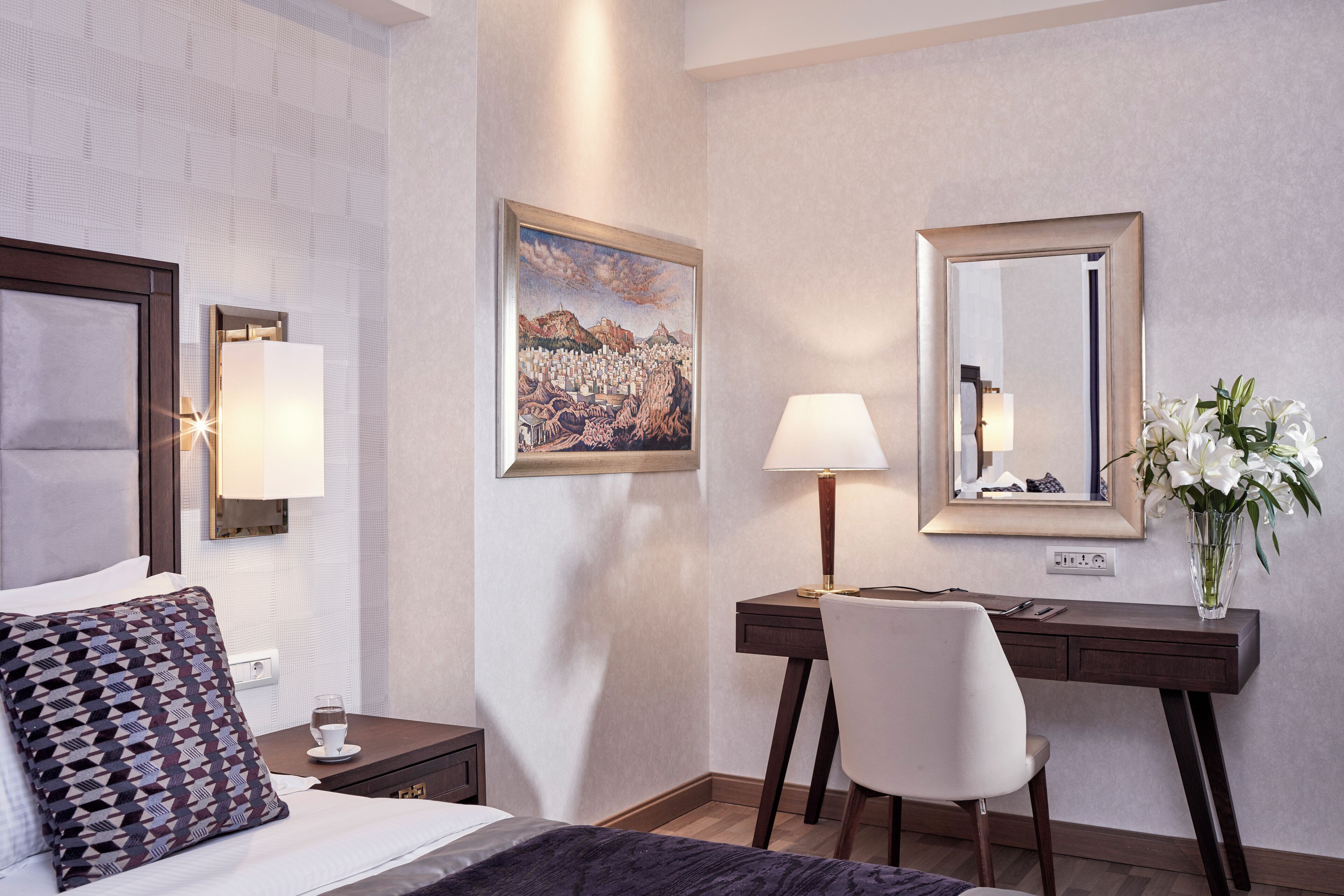 A hotel room with a bed, a desk, a chair, a lamp, a vase with flowers, a painting, and a mirror.