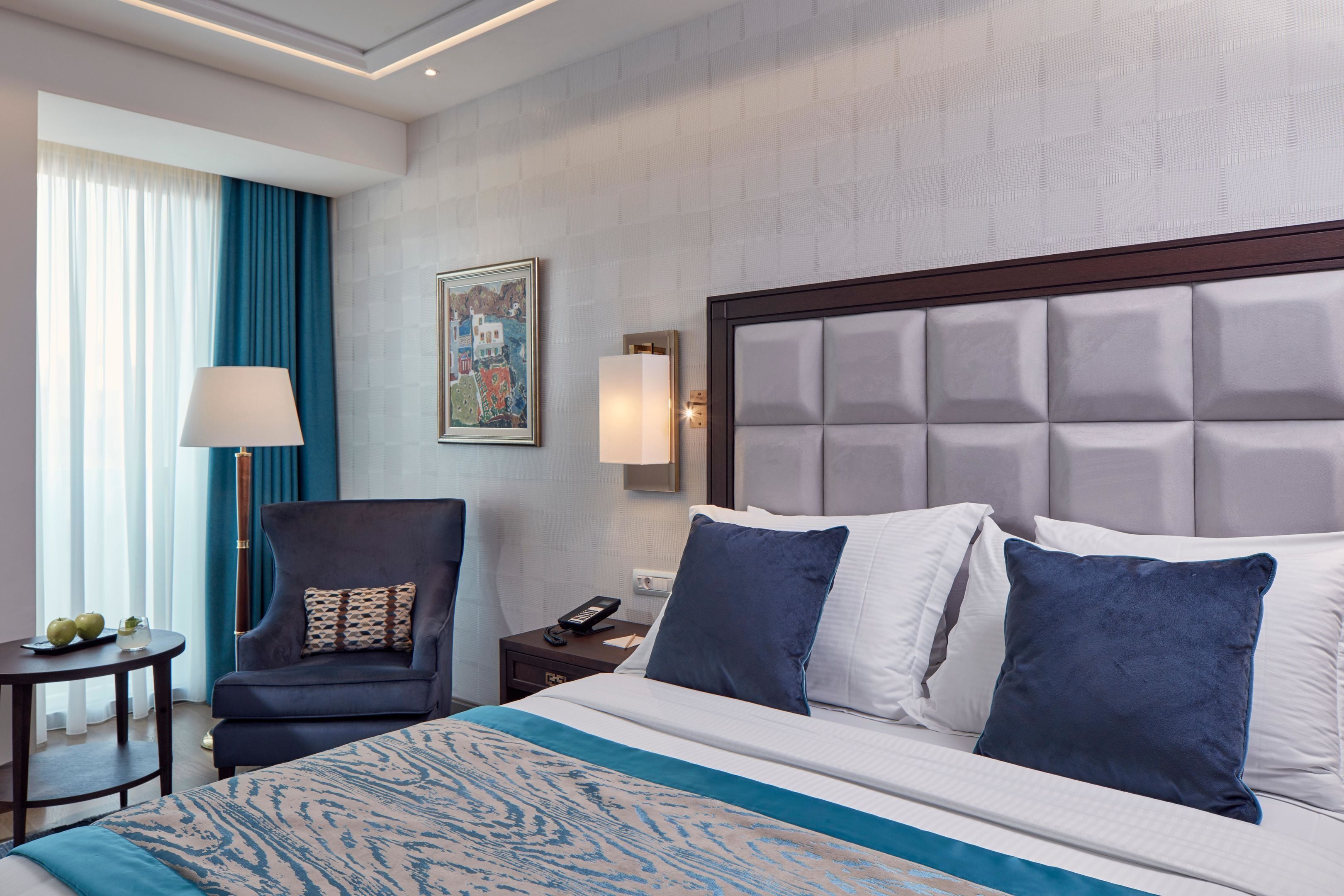 A hotel room with a bed, a chair, a nightstand, a lamp, and a painting on the wall.