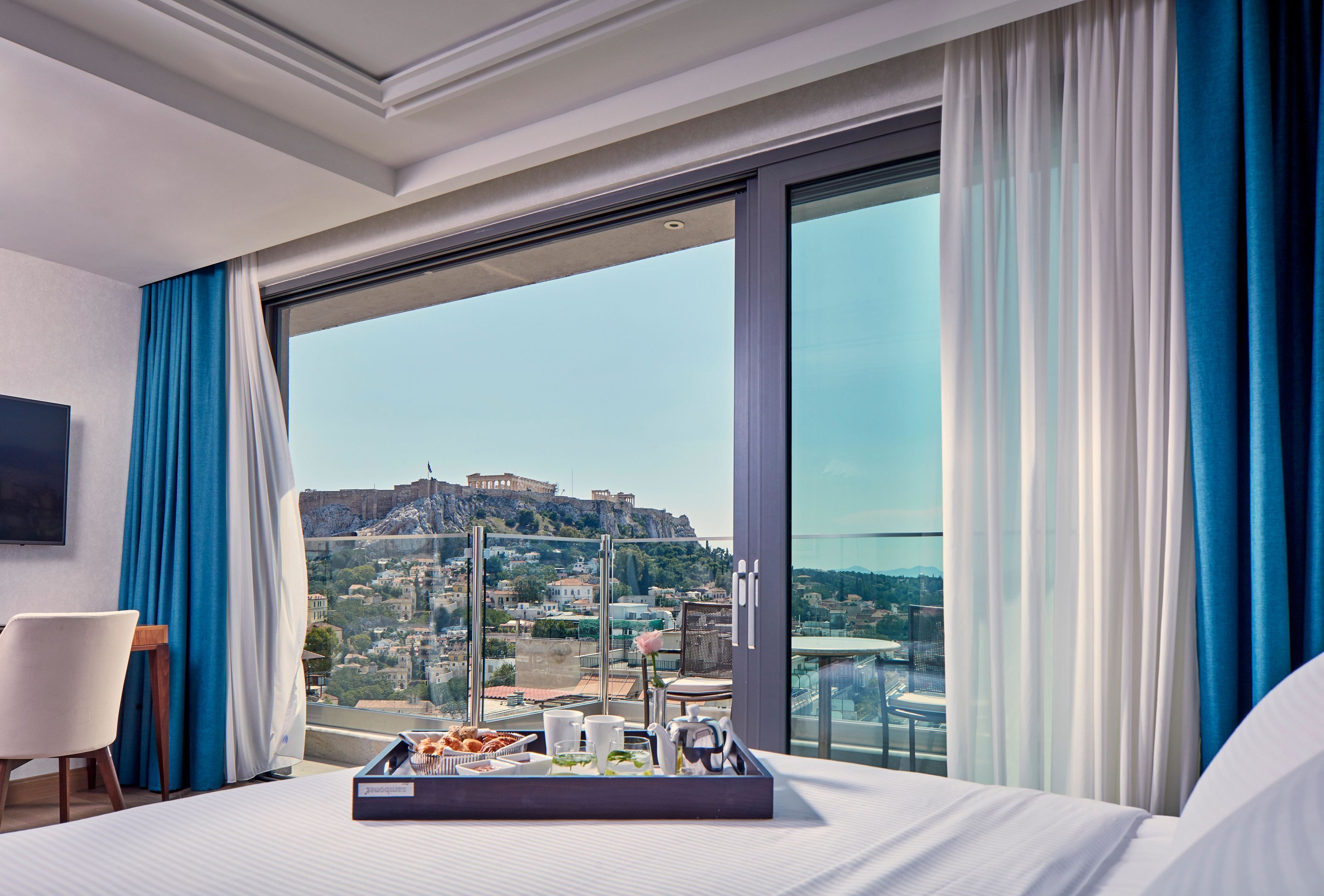 Une chambre d’hôtel avec une grande fenêtre, un lit, un plateau avec des articles pour le petit-déjeuner et une vue sur un paysage urbain comprenant un bâtiment historique.