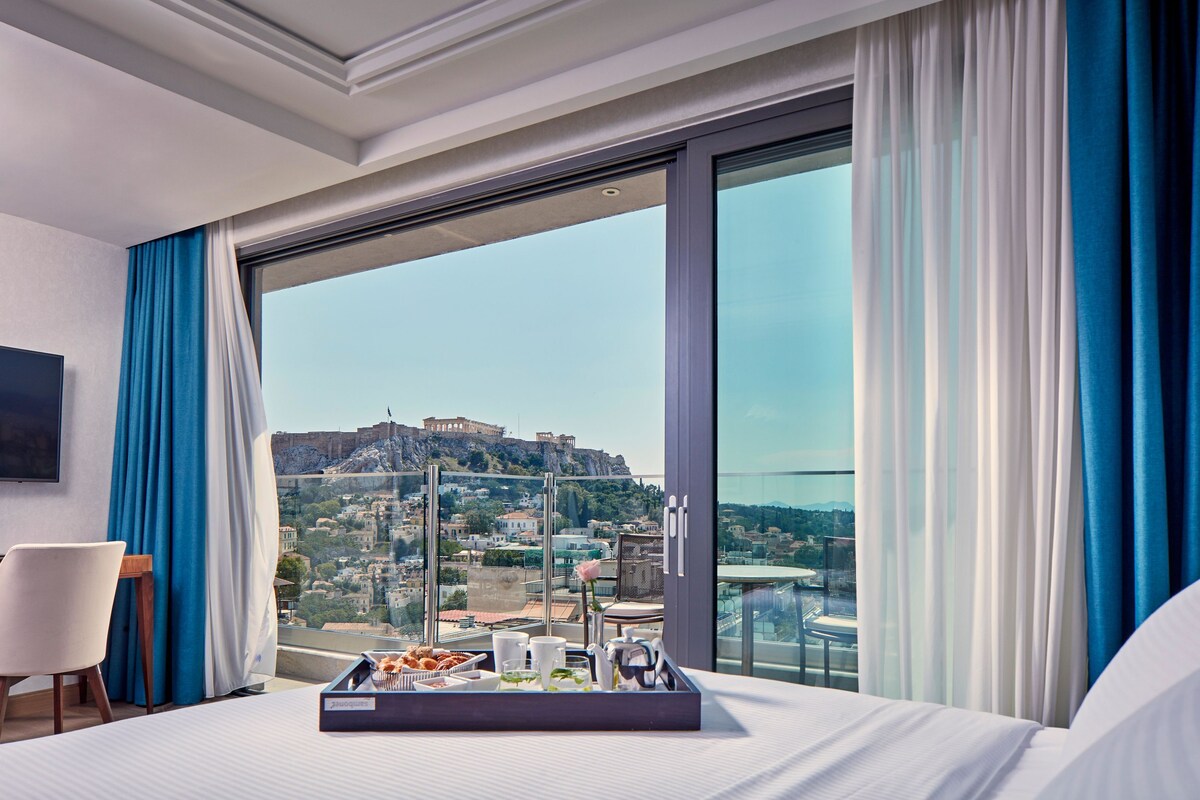 A hotel room with a large window, a bed, a tray with breakfast items, and a view of a cityscape with a historic building.
