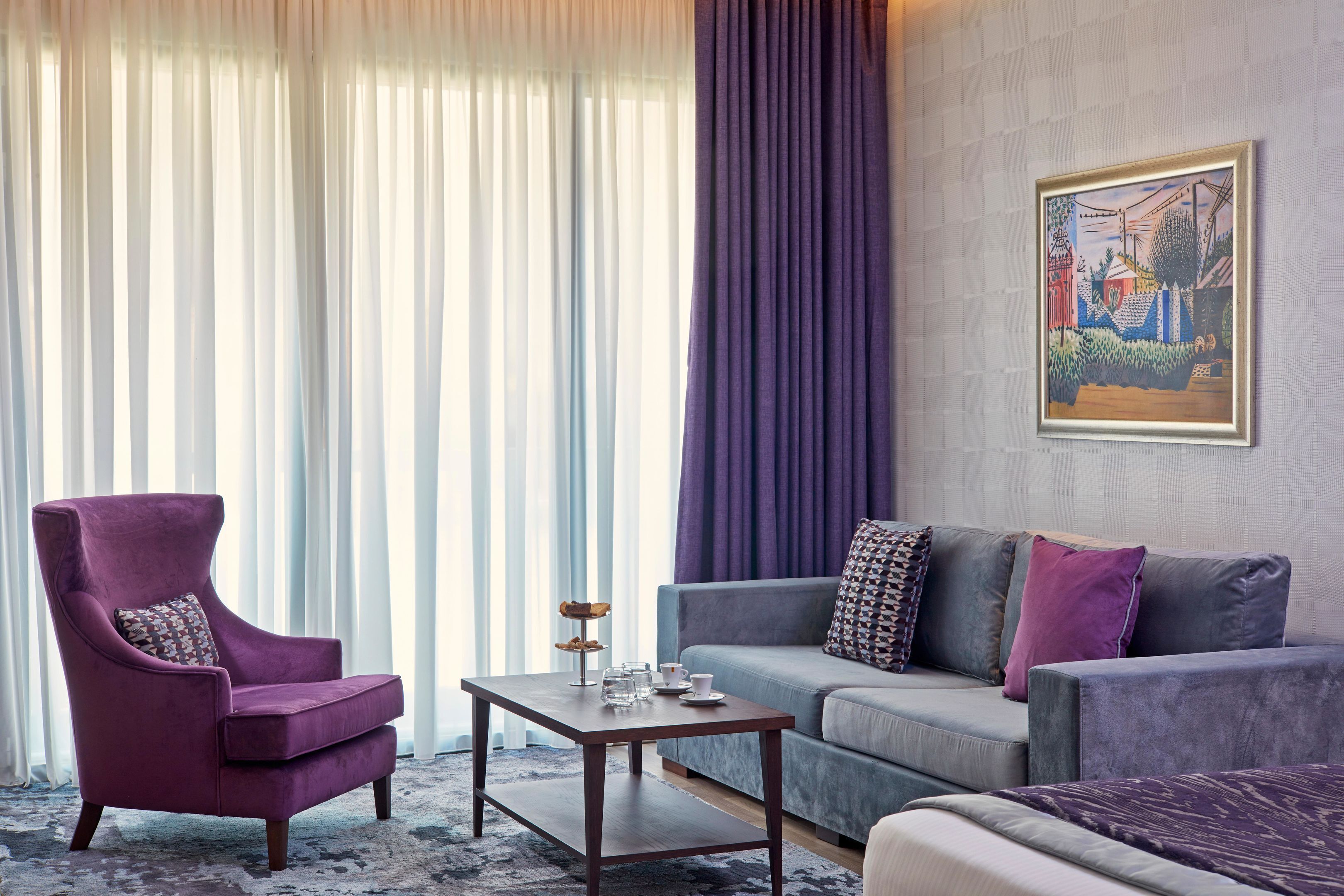 A modern living room with a purple armchair, a grey sofa, and a coffee table with a tray of pastries.