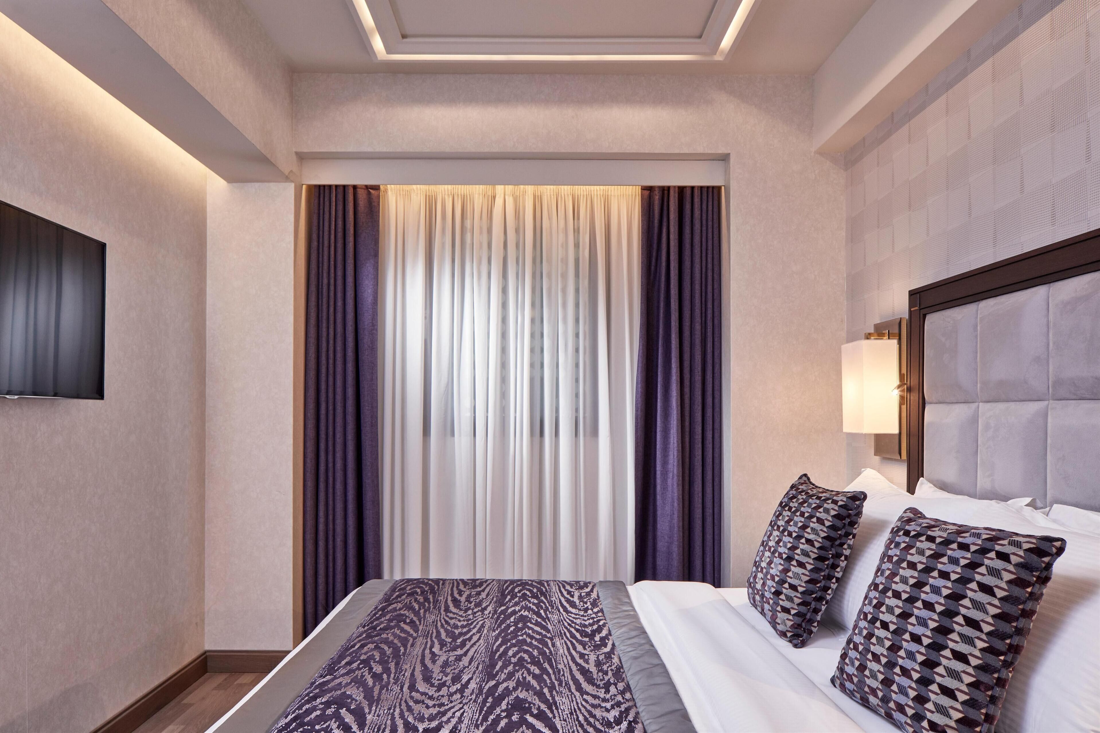 A hotel room with a large bed, a flat-screen TV mounted on the wall, and a window with curtains.
