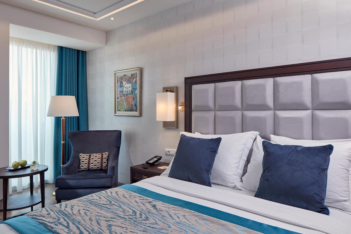 A hotel room with a large bed, a chair, a nightstand, a lamp, and a painting on the wall.