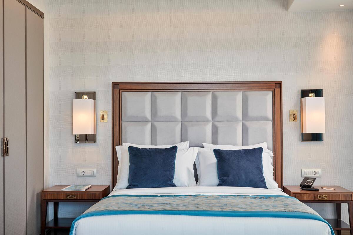 A hotel room with a tufted headboard, two bedside tables with lamps, a bed with a blue and white bedspread, and a wall-mounted telephone.