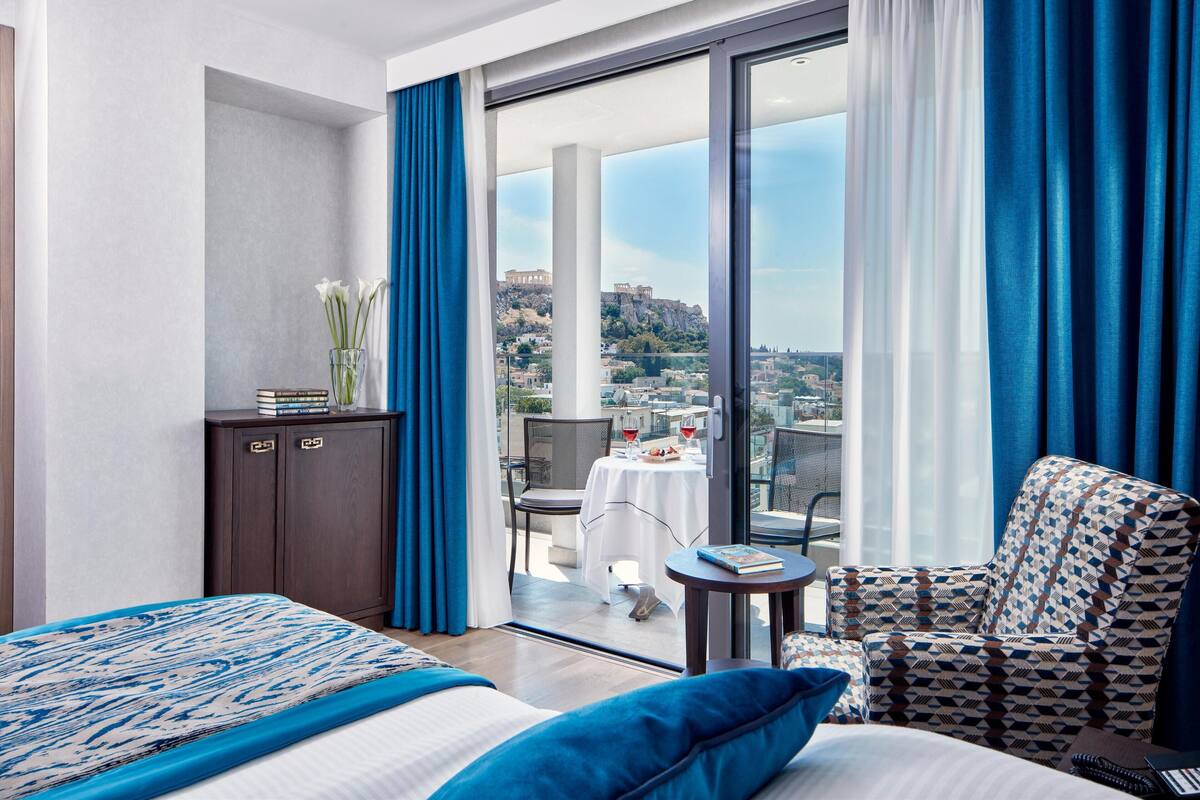 A hotel room with a bed, a desk, a chair, and a balcony with a view of a cityscape.