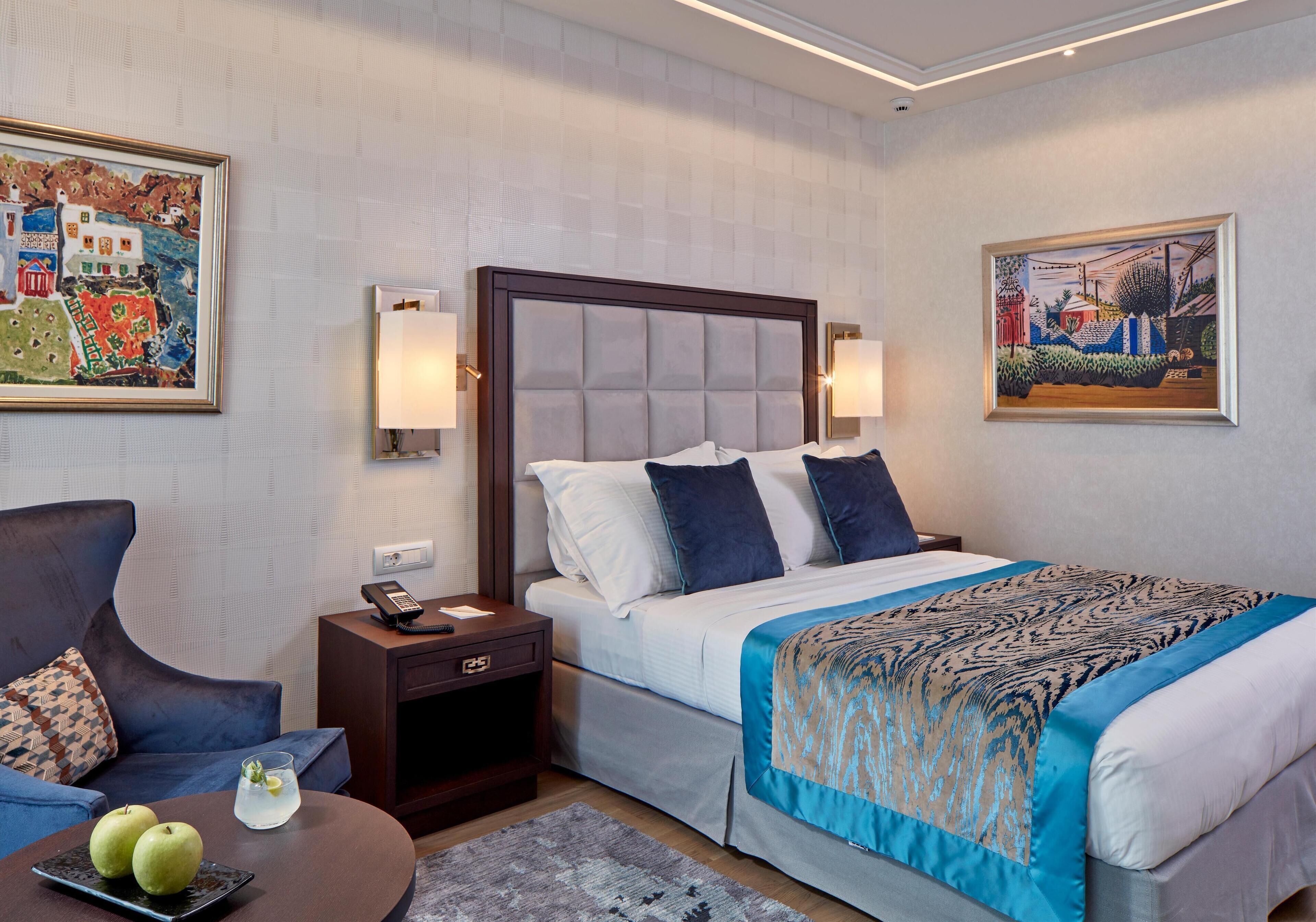 A hotel room with a bed, a chair, a small table with apples, and two paintings on the walls.