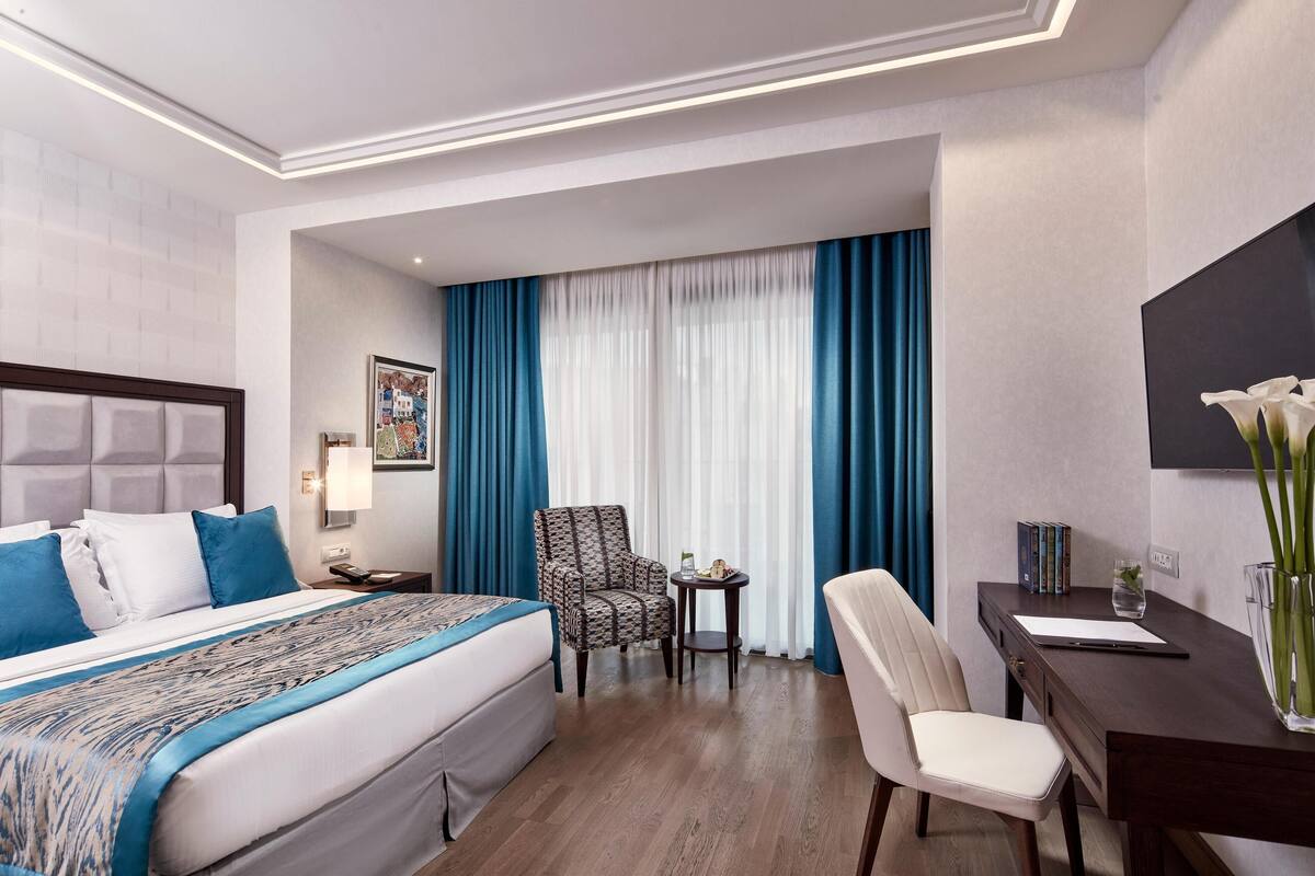 A hotel room with a bed, a desk, a chair, a small table, and a window with curtains.