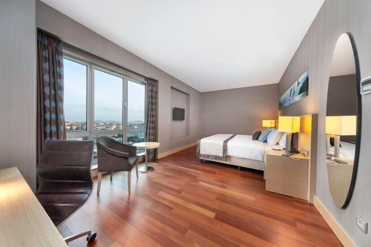 Habitación de hotel con cama, mesitas de noche, escritorio, silla, una pequeña mesa y un ventanal con vistas a la ciudad.