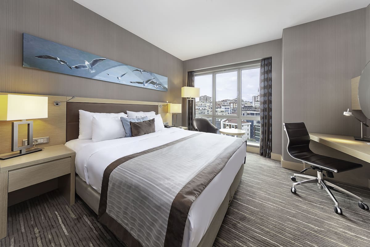 A hotel room with a large bed, a desk, a chair, and a view of the city.