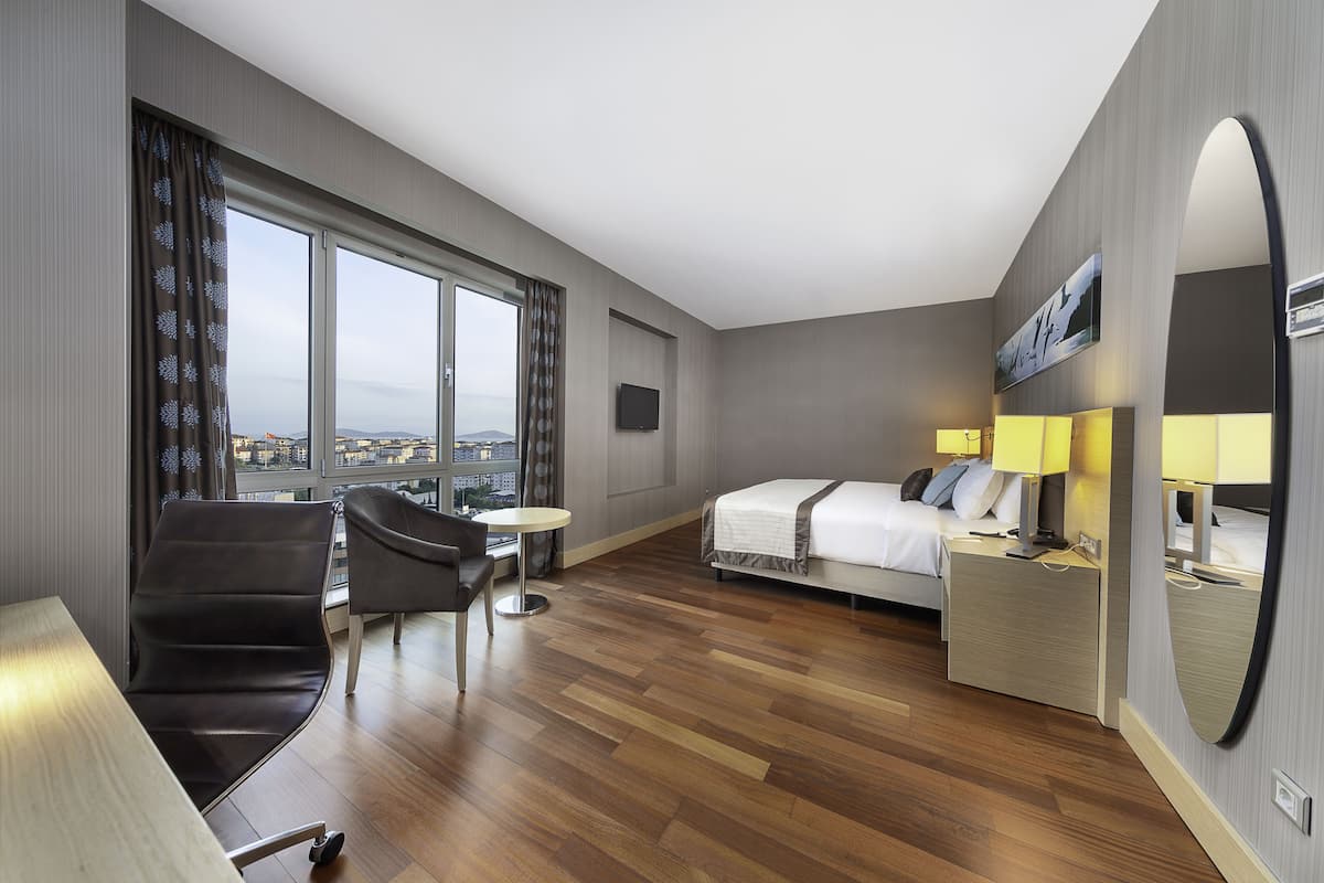 A hotel room with a bed, bedside table with lamps, a chair, a small round table, and a large window offering a city view.