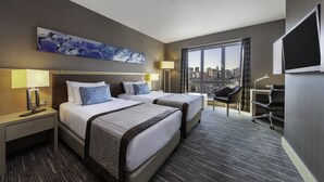 Habitación de hotel con dos camas, un escritorio, una silla, un televisor y un ventanal con vistas a la ciudad.