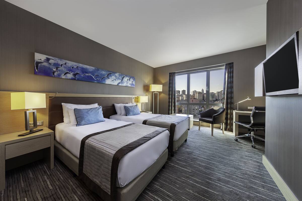 A hotel room with two beds, a desk, a chair, a TV, and a large window with a city view.