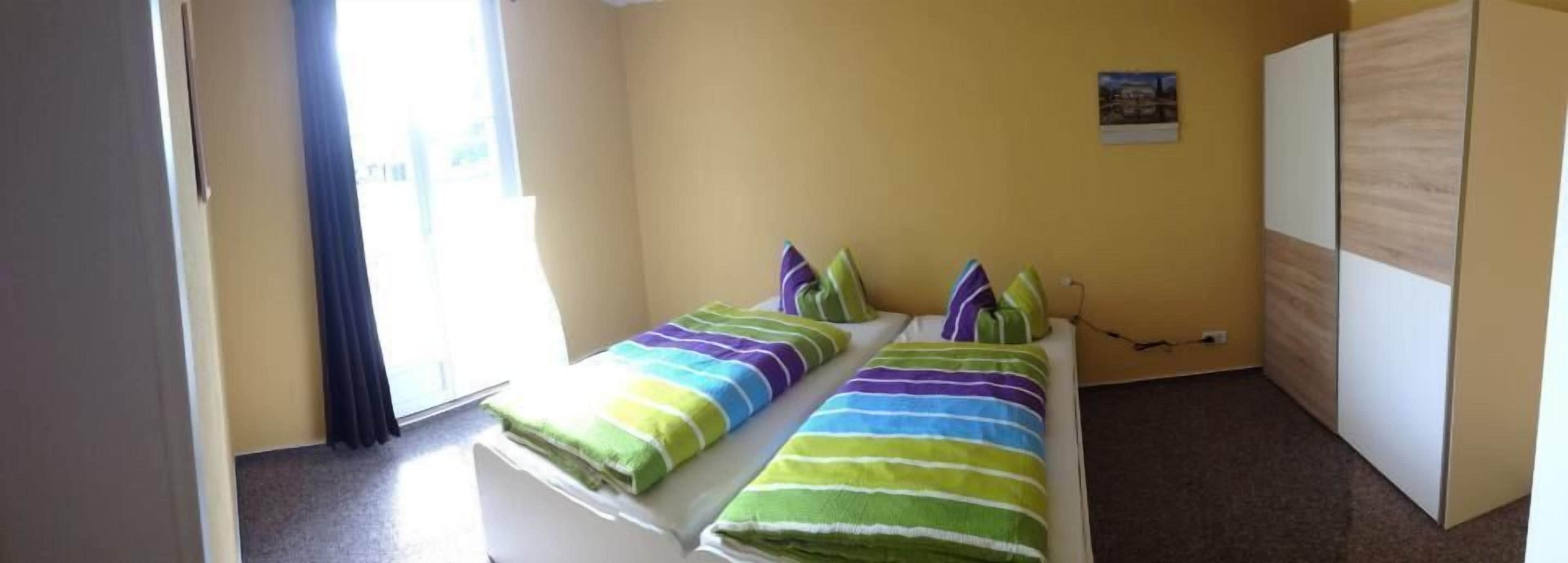 A small, single-bed room with a plain yellow wall, a window with curtains, and a white wardrobe.