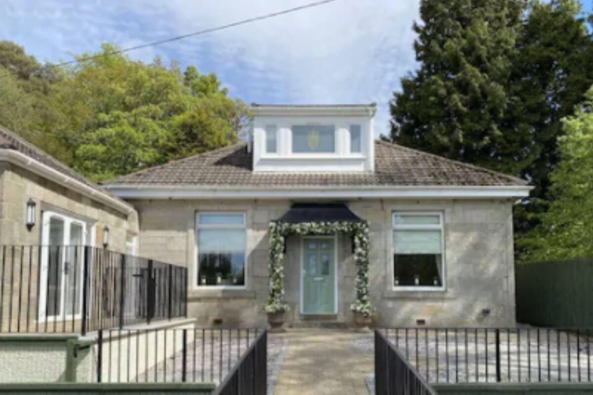 A two-story house with a tiled roof, a front porch with a green door, and a small garden area.