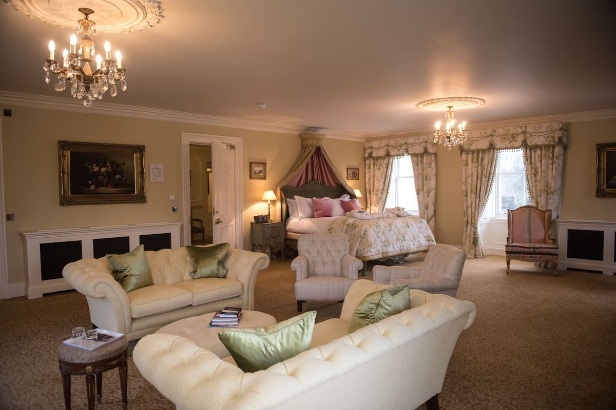 A spacious room with a large bed, two armchairs, a chaise lounge, and a chandelier.