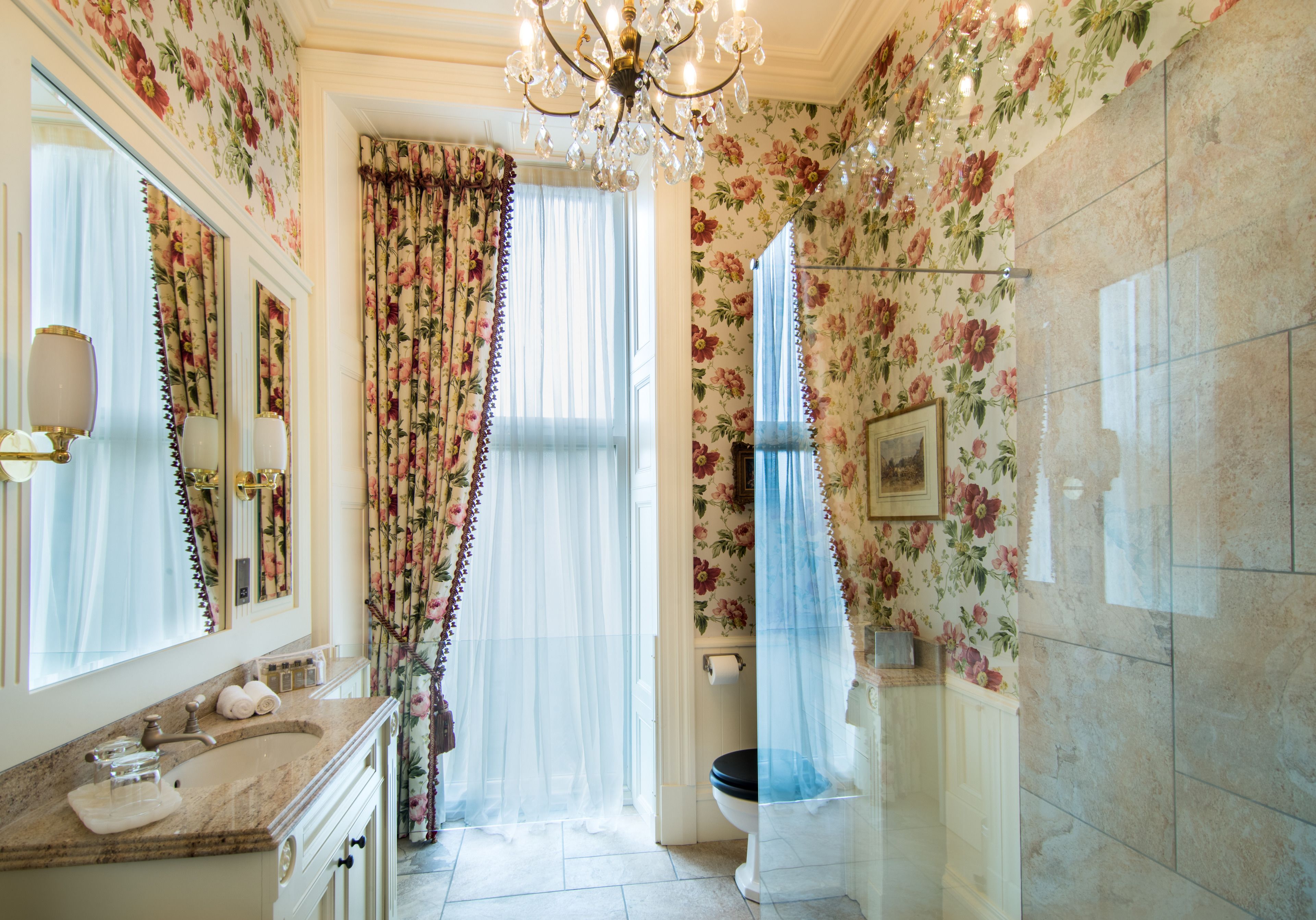 A bathroom with floral wallpaper, a chandelier, a sink with a marble countertop, and a toilet with a blue shower curtain.