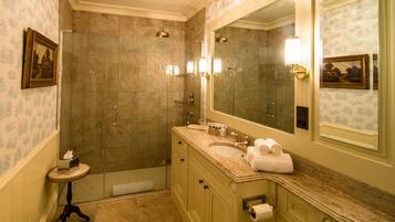 A bathroom with a shower, sink, and large mirror. There is a towel rack and a small table with a towel.