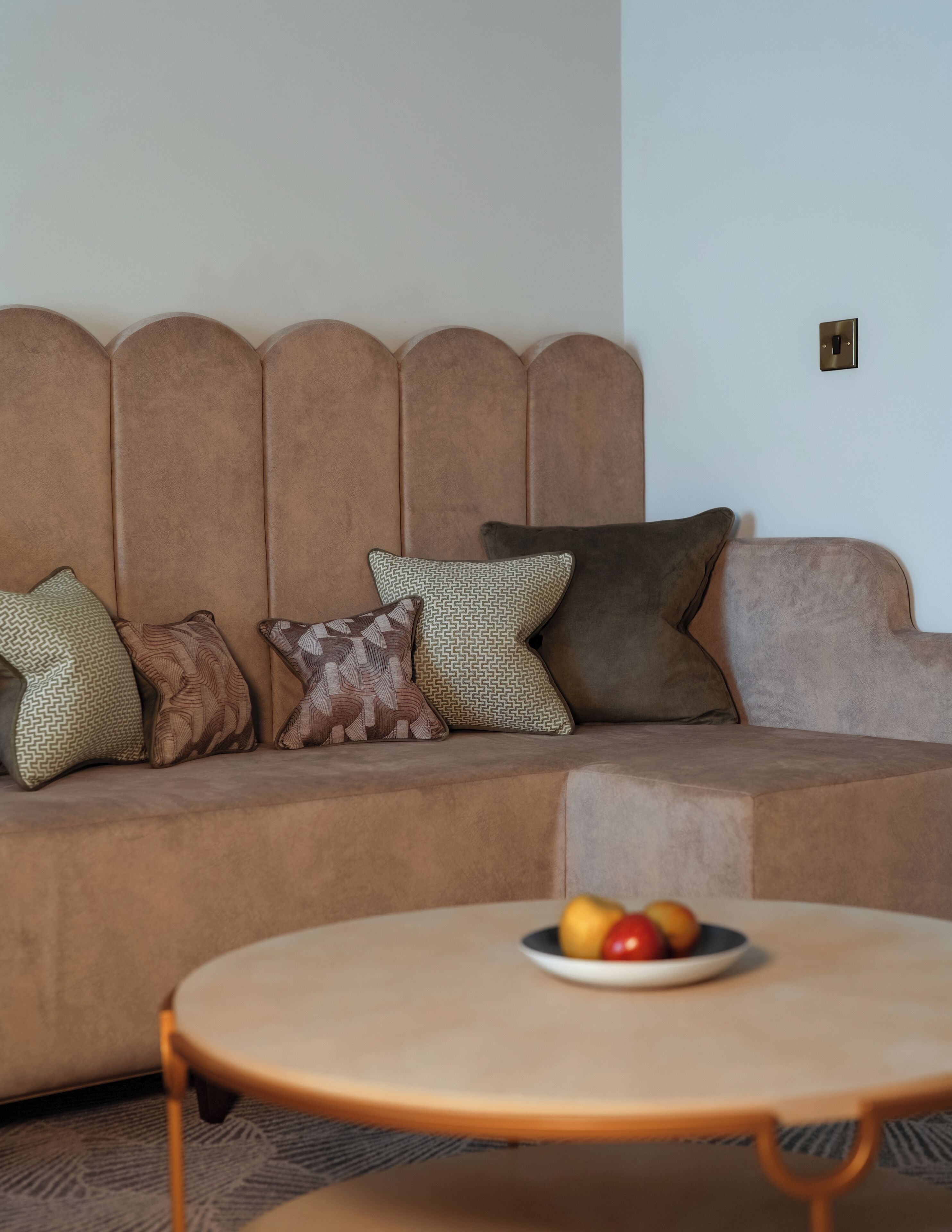 Ein Wohnzimmer mit einem braunen Sofa, gemusterten Kissen und einem runden Couchtisch mit Früchten.