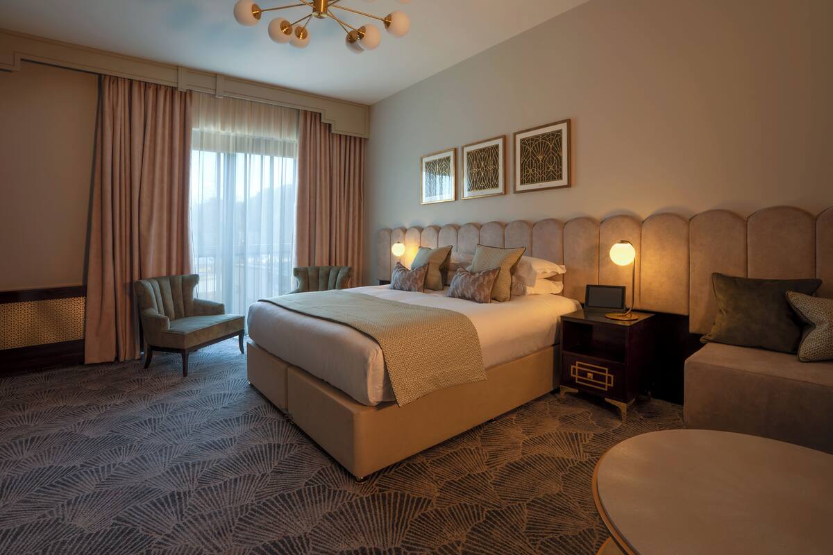 A hotel room with a large bed, a bedside table with a lamp, a chair, a round table, and a view of the outdoors through the window.